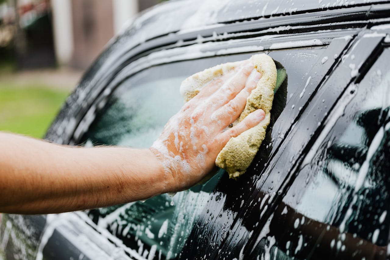 A hand scrubs a car window with a sponge, covered in soap suds, during a car wash.